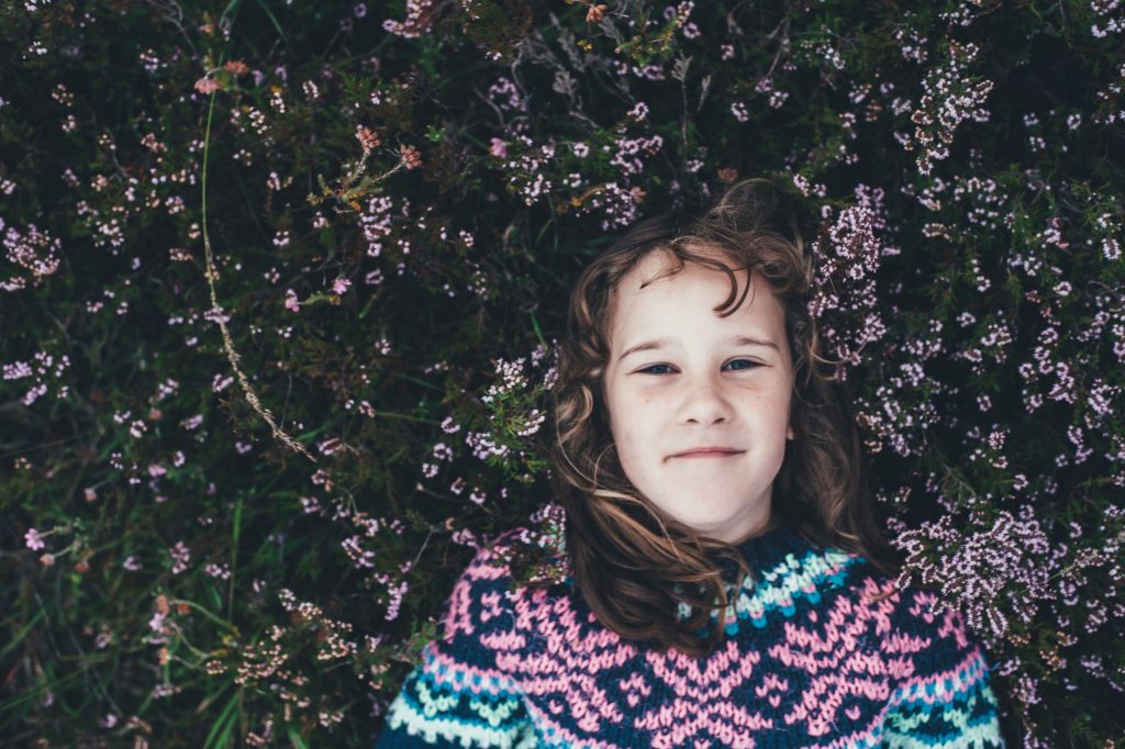A young girl wearing a woolly jumper laying in a field of purple flowers.