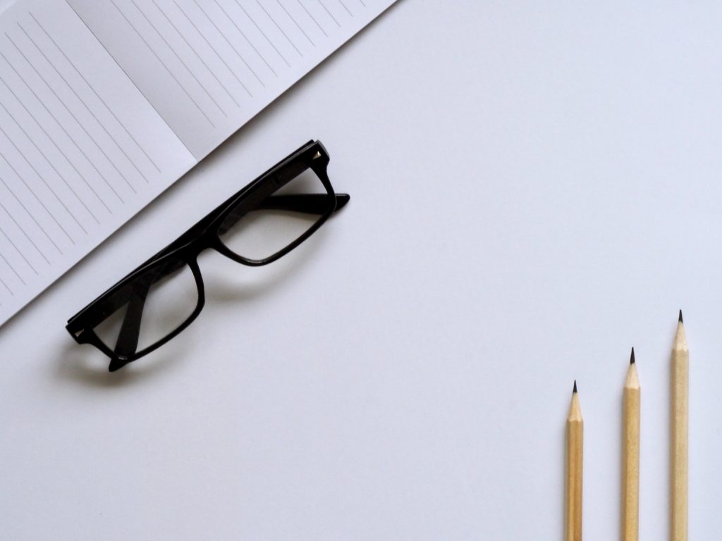 A pair of glasses, three pencils and a notebook against a white background.