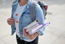 A young woman in a denim jacket wearing a back pack and holding a stack of folders and books.