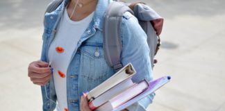 A young woman in a denim jacket wearing a back pack and holding a stack of folders and books.