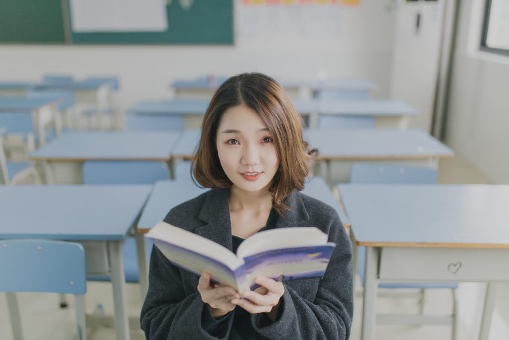 A female teacher standing at the front of a classroom holding an open book.