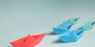 A red paper boat followed by three blue paper boats against a blue background.